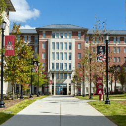 Students walking and on bikes walk on a brick courtyard.  Behind them is a six story brick residence hall flanked by trees.