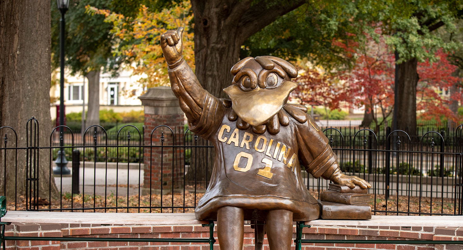 The bronze Cocky statue sits on a bench in front of a wrought iron fence with trees with fall leaves in the background.