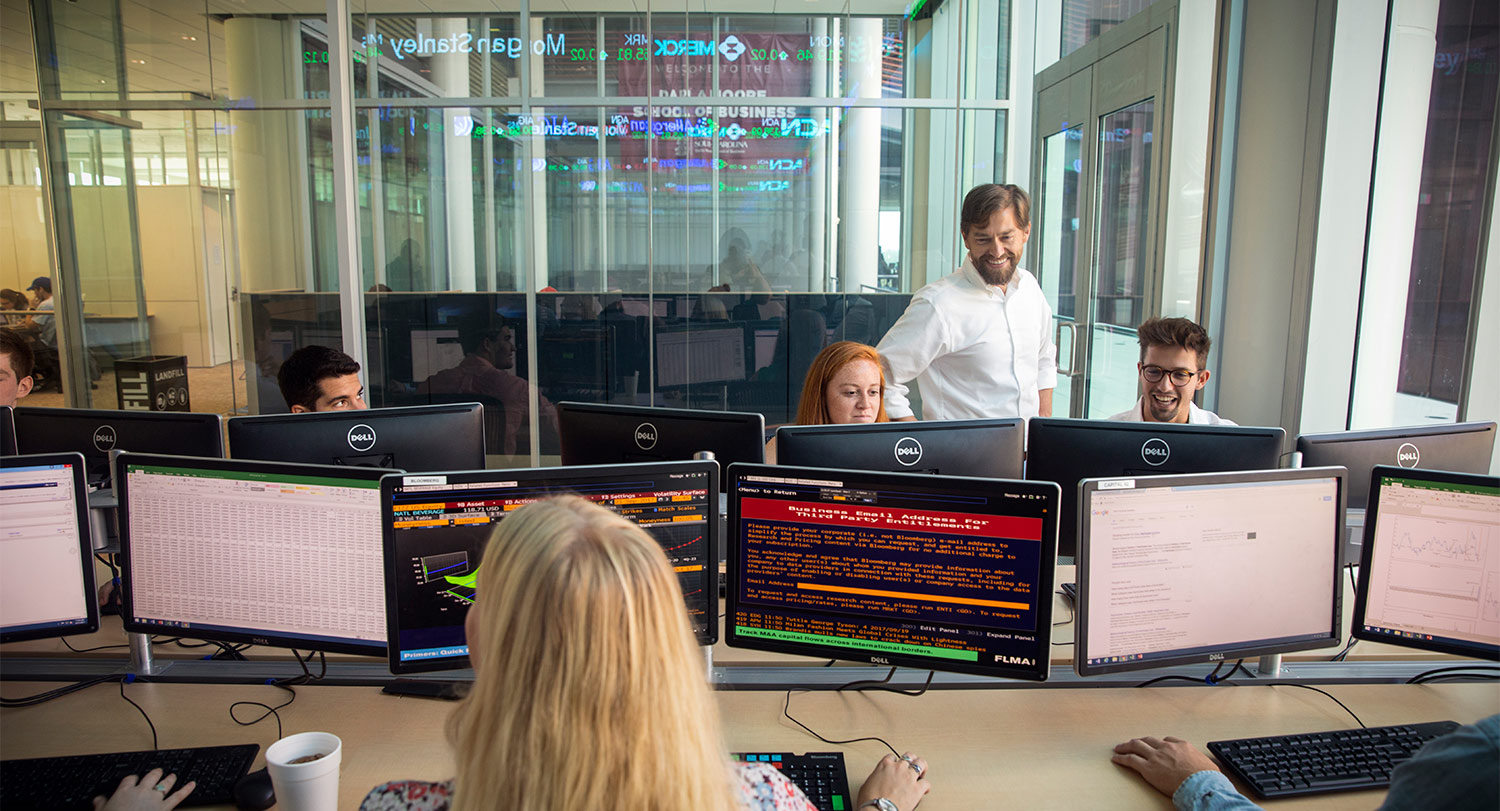 A business class in a computer lab with the professor standing with two students looking at a monitor. 