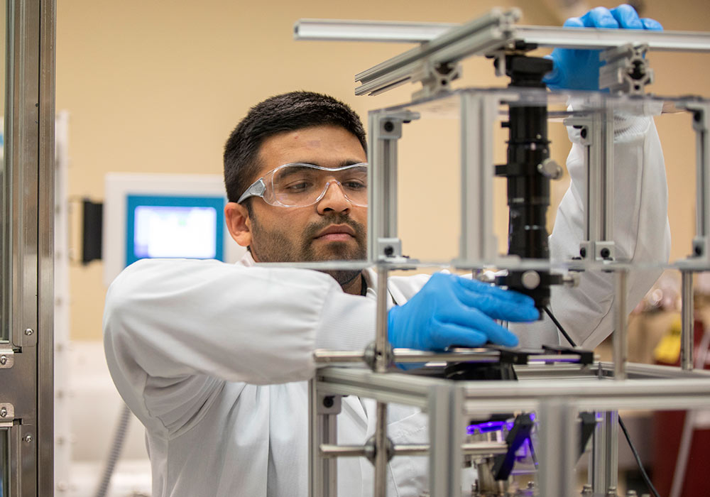 Graduate student wearing a lab coat works on a piece of equipment.