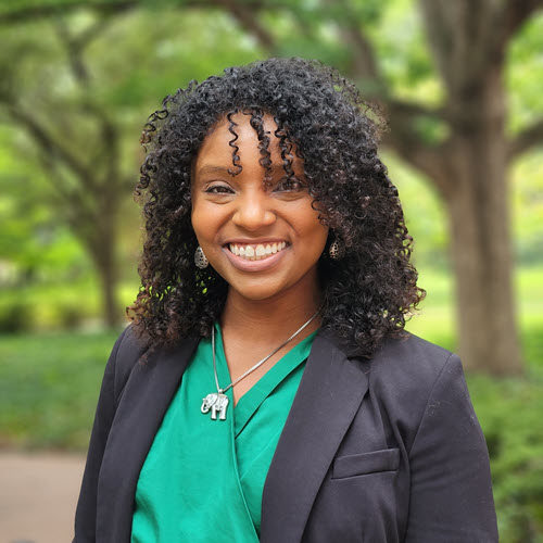 Michelle Brown headshot from mid torso. She has black, curly hair and is wearing a gray blazer and a navy blue button up blouse with a silver necklace.