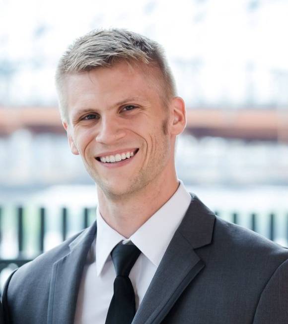 Stephen Taylor is smiling at the camera, wearing a gray suit and a black tie. He is posed in front of a blurred skyline.