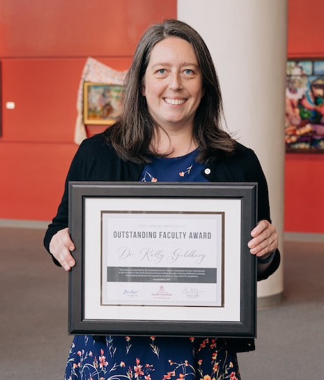 Dr. Kelly Goldberg standing in the Koger Center lobby holding her 2024 SCHC outstanding faculty award